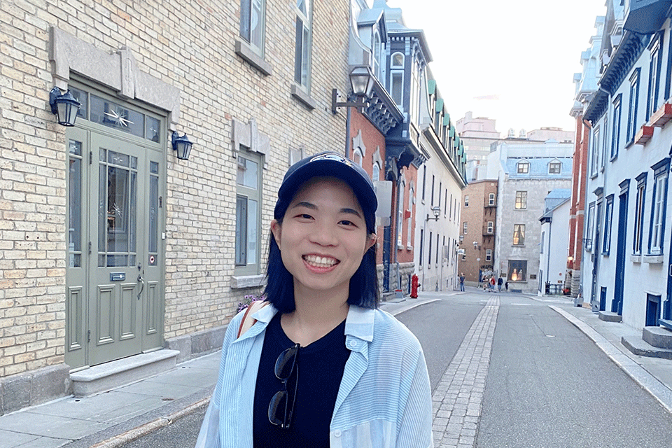 一名女子(冉冉升起的明星Irene Wang)穿着黑色上衣，白色敞开的衬衫，微笑着站在两旁都是房屋的街道上。