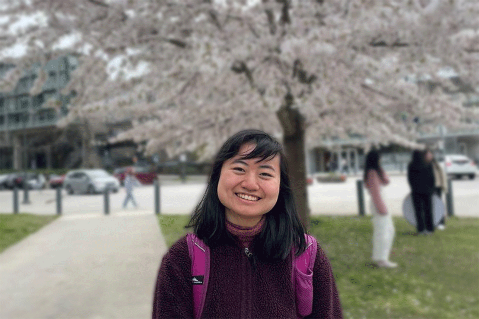 一名女子(冉冉升起的新星张菲比)身穿黑色连帽衫，微笑着站在樱花树前。