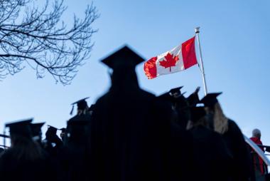 蓝色的天空中飘扬着加拿大国旗，前景中是UBC 2022届毕业班的影子，他们戴着帽子，穿着长袍。开云体育最新网站